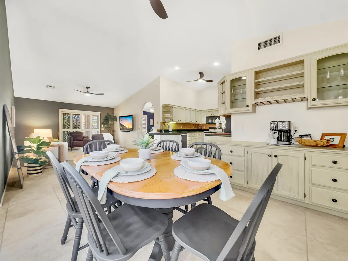 An open-plan area where a round wooden dining table sits adjacent to the living room and kitchen spaces. The scene is accented by greenery near a window, contemporary wall art, and plenty of natural light, creating an inviting setting for meals, conversation, and shared experiences.