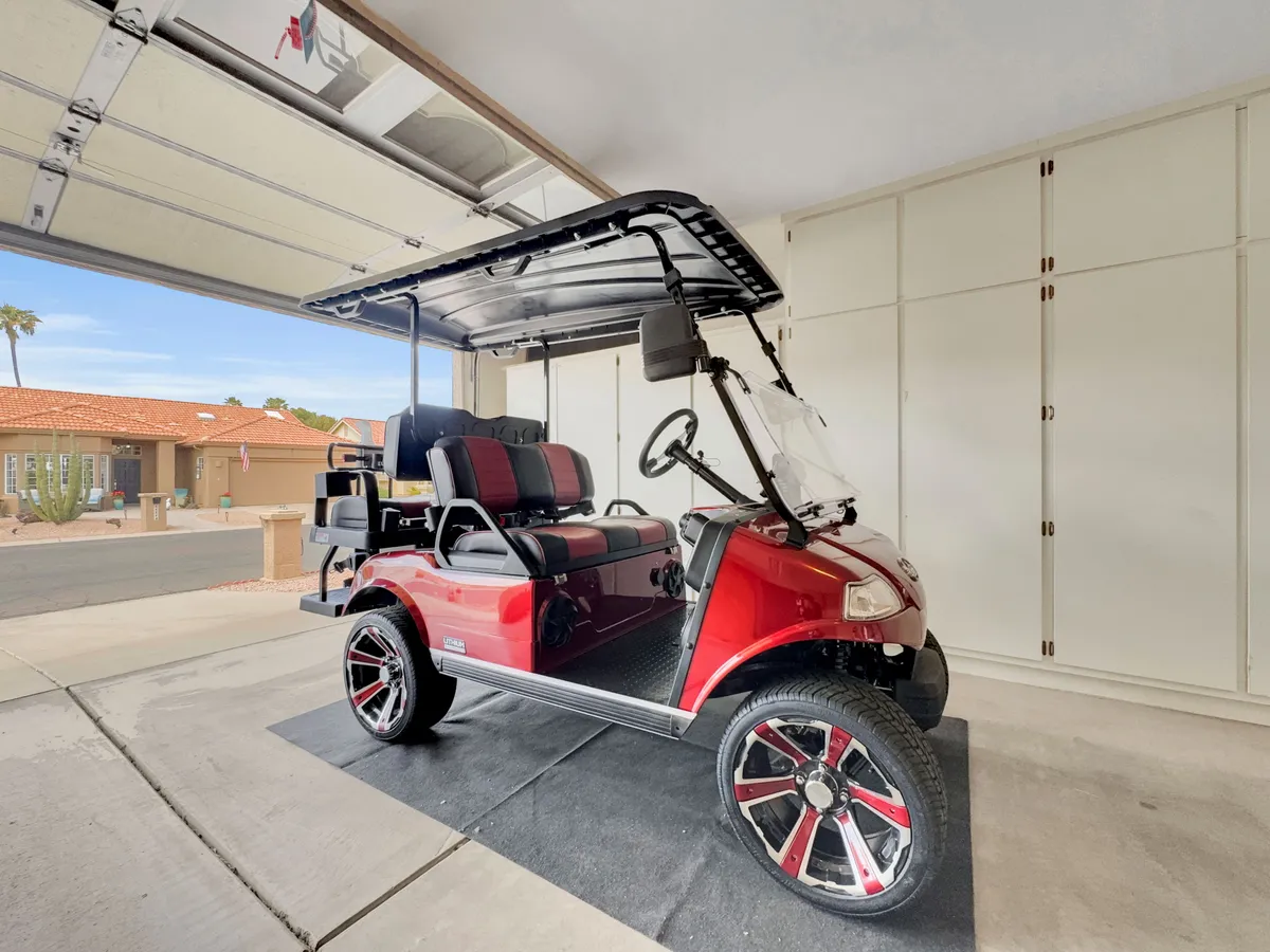 A red golf cart is parked in an open garage, with storage cabinets along the walls. The scene emphasizes leisure and convenience for getting around the community.
