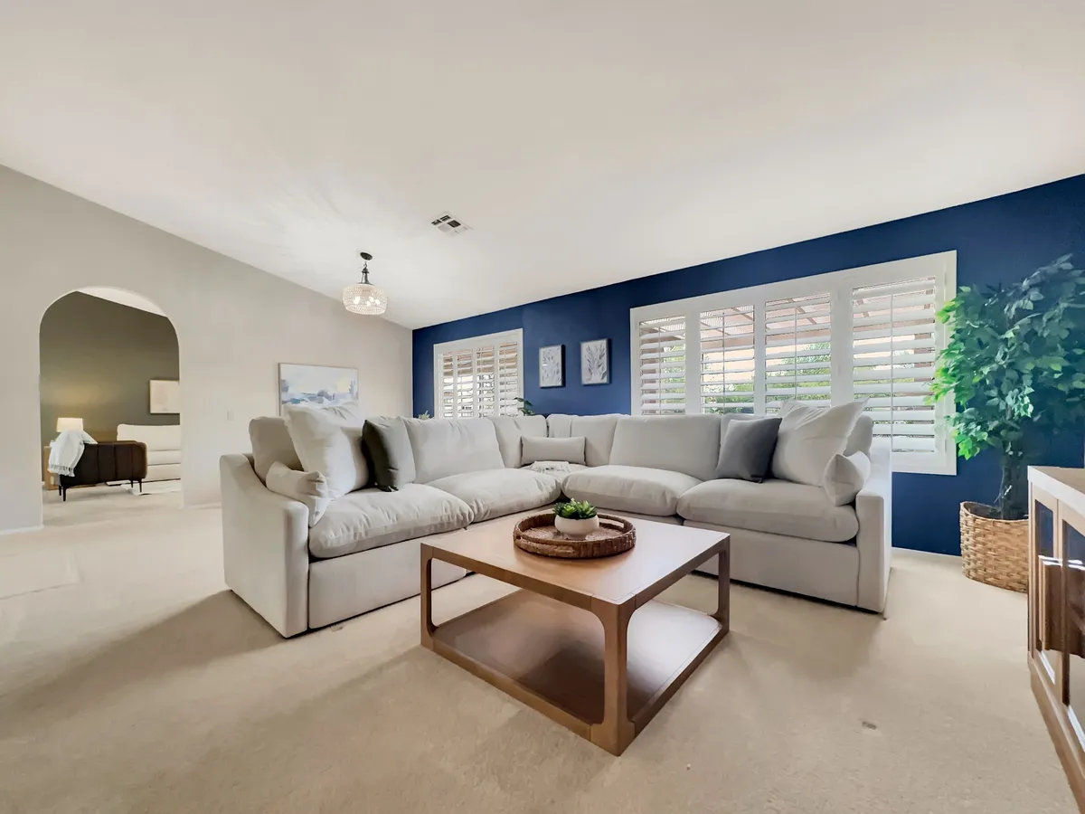 A spacious living room featuring a large sectional sofa, a wooden coffee table, decorative wall art, expansive windows with shutters, and a lush houseplant. Sunlight streams in through the windows, illuminating the cozy seating area and creating a cheerful, welcoming ambiance.