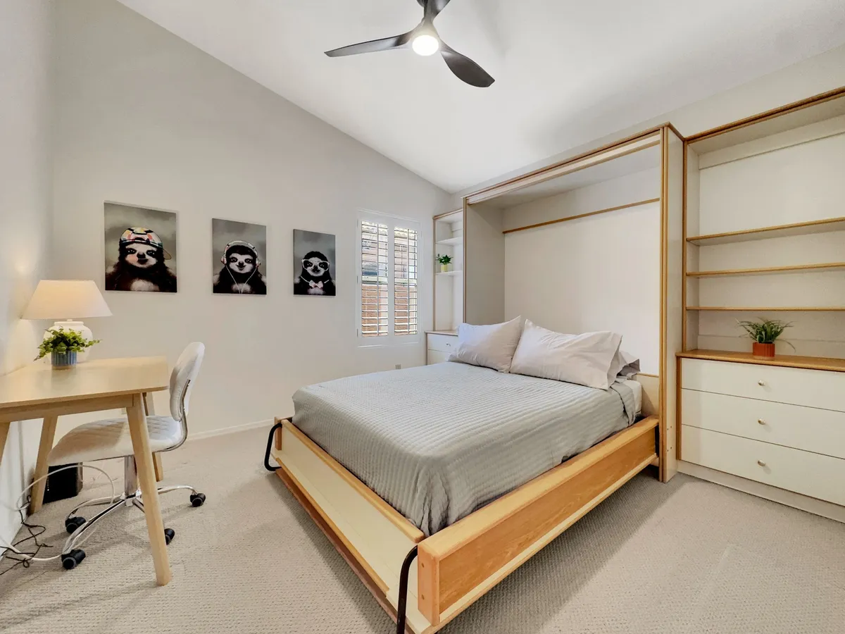 A cozy bedroom with soft gray bedding on the bed, a desk with a plant and lamp, and playful sloth artwork on the wall. The space combines comfort for sleeping with a functional workspace, illustrating versatility for guests or remote workers.