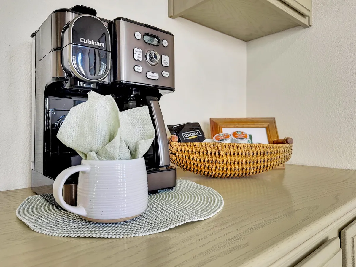 A close-up of a coffee maker on a countertop, accompanied by a ceramic mug and woven basket. The arrangement evokes the comfort of brewing fresh coffee and enjoying simple morning rituals in the kitchen.